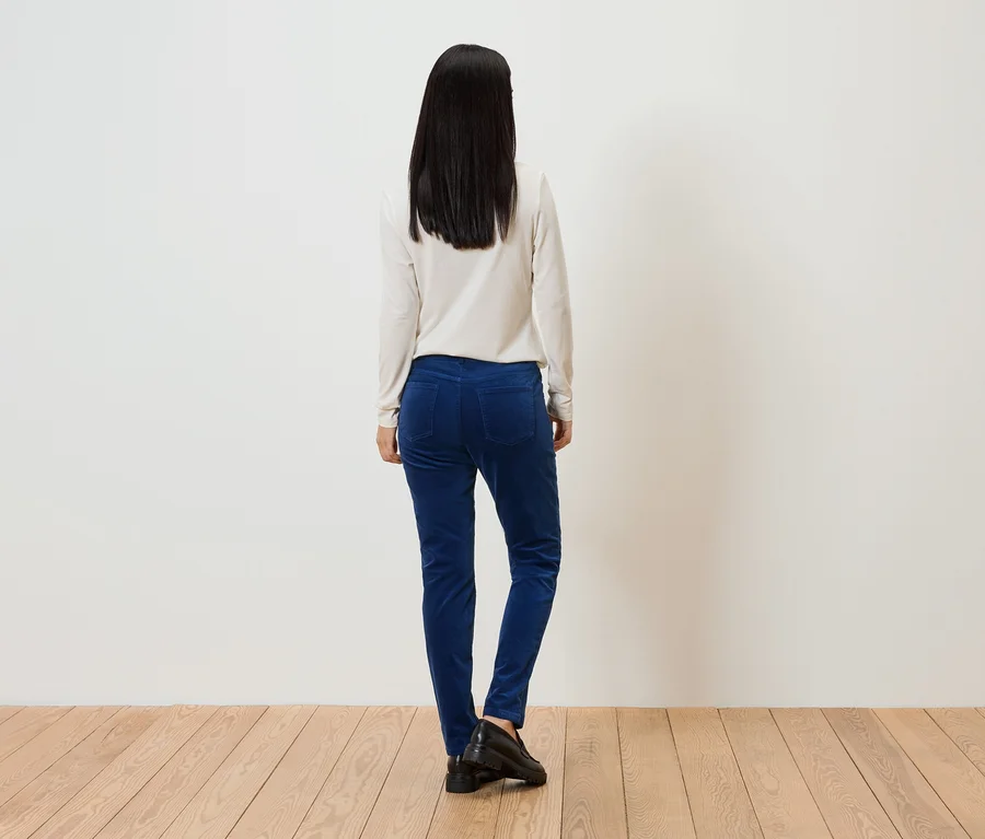 Eine Frau mit langen, dunklen Haaren steht mit dem Rücken zum Betrachter, trägt ein helles Oberteil, blaue Hosen und schwarze Schuhe.