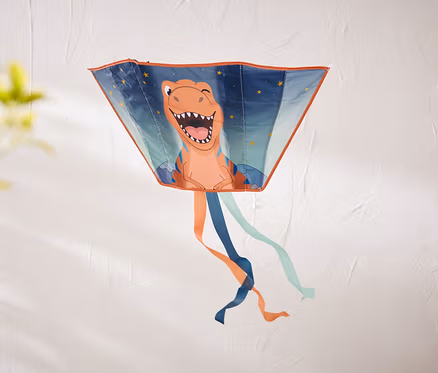 Ein trapezförmiger Drachen mit einem lachenden orangefarbenen Dinosaurier und Sternen hängt an einer strukturierten weißen Wand. Unten hängen orange, dunkelblaue und hellblaue Bänder.