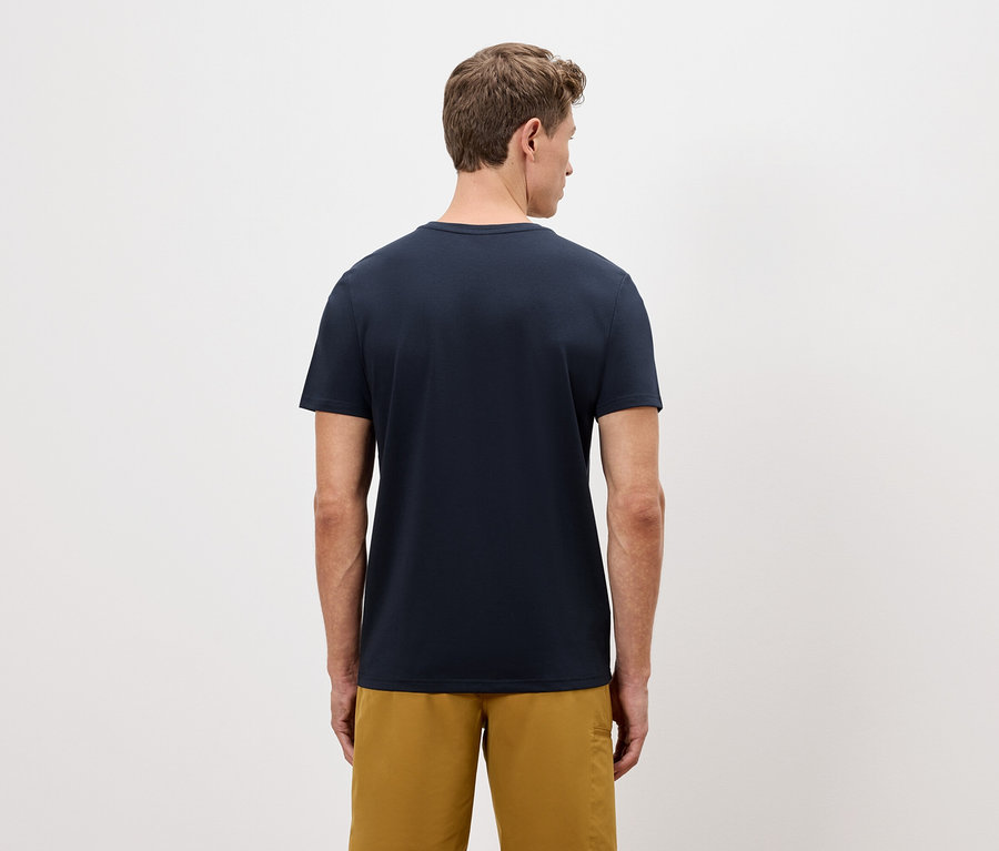 Ein Mann von hinten, der ein marineblaues T-Shirt und goldene Shorts vor einem einfachen Hintergrund trägt.