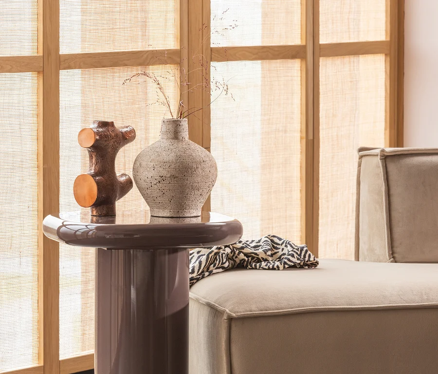 Ein brauner Beistelltisch mit einer strukturierten hellen Vase und einer metallischen Skulptur. Rechts daneben steht ein beiges Sofa mit einem schwarz-weiß gemusterten Überwurf vor einem Fenster mit Holzrahmen und gewebten Jalousien.