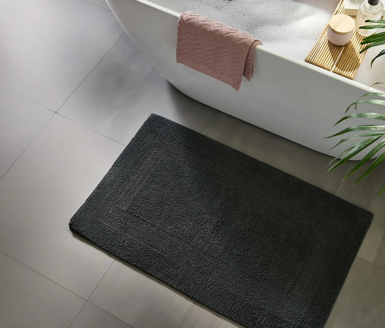 Dunkelgraue Badematte auf hellgrauen Fliesen vor einer weißen Badewanne mit rosa Handtuch, Badeschaum, einem Holzbadebrett und einer Pflanze.