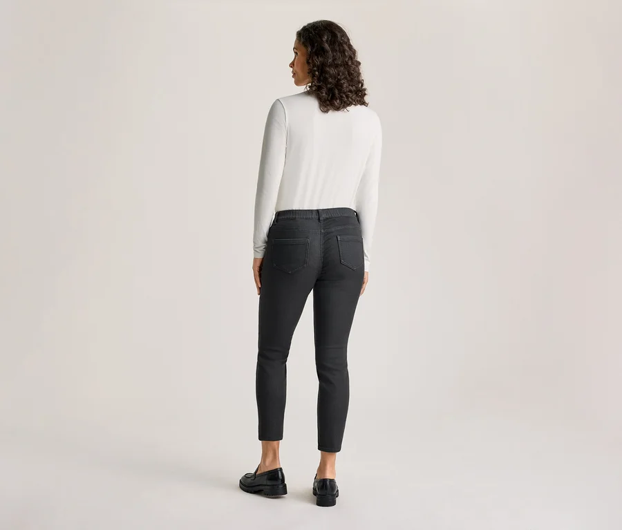 Eine Frau steht mit dem Rücken zum Betrachter und blickt leicht nach links. Sie trägt ein weißes Langarmshirt, dunkelgraue Treggings - Fit »Nelly« und schwarze Loafer.