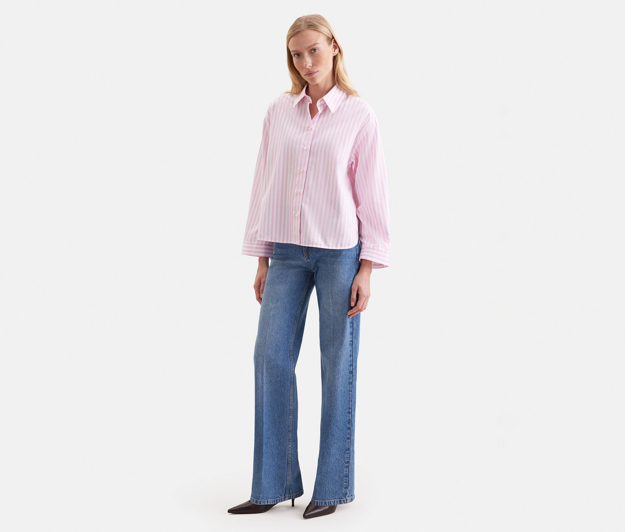 Frau steht, trägt eine rosafarbene Seidensticker Oversize Hemdbluse mit Streifen und blaue Jeans.