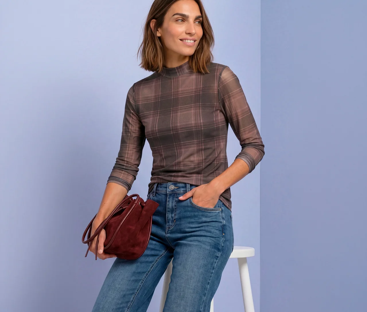 Eine lächelnde Frau sitzt auf einem Hocker und trägt ein kariertes Street One Mesh-Shirt, blaue Jeans und hält eine bordeauxrote Beuteltasche.