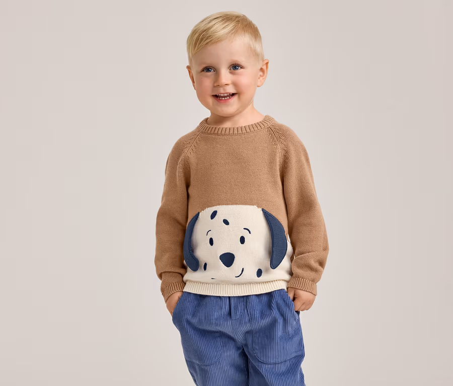 Ein lächelnder kleiner Junge mit blondem Haar steht mit den Händen in den Taschen und trägt einen braunen Pullover mit einem weißen und blauen Hundegesicht auf der Vorderseite sowie eine blaue Cordhose.