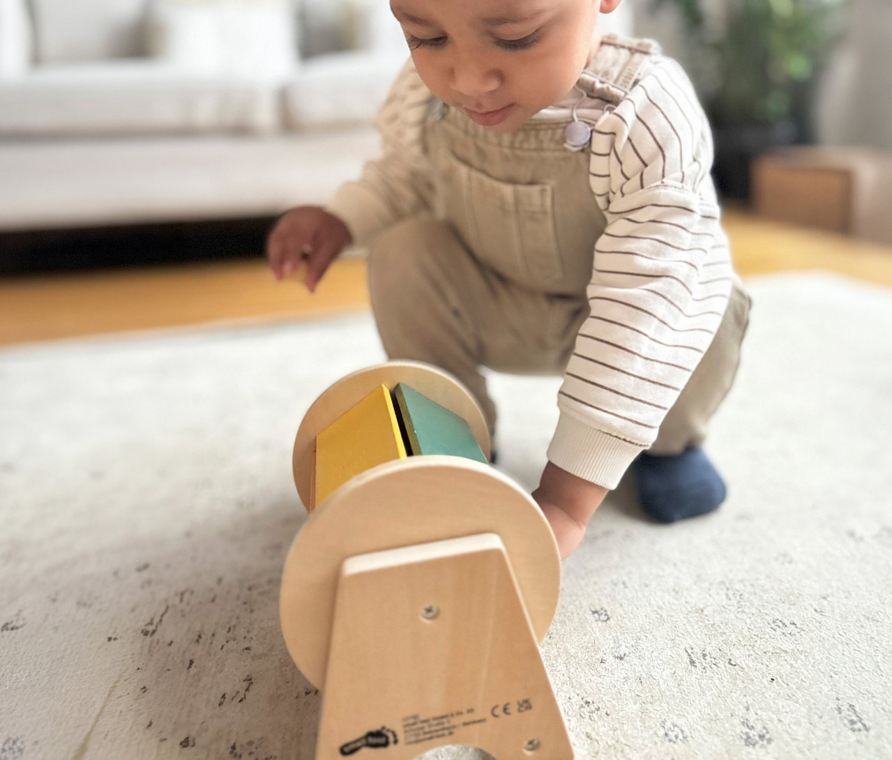 Ein Baby in beiger Latzhose und gestreiftem Shirt hockt auf einem Teppich und blickt auf ein rollendes Holzspielzeug aus dem small foot Montessori-Bundle 3 mit gelben und grünen Blöcken.