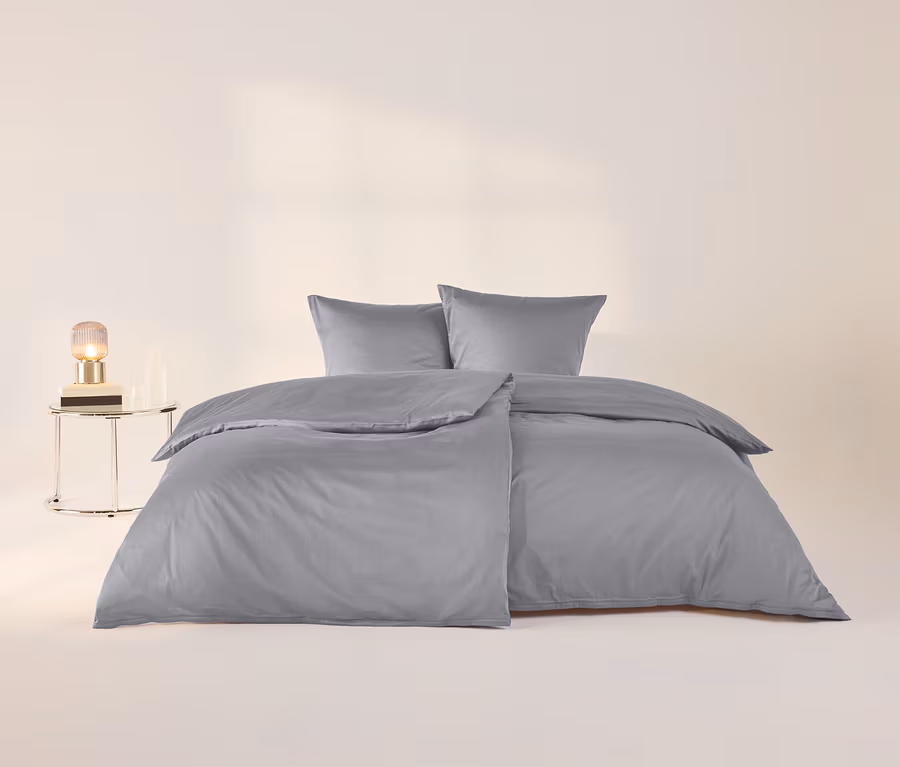 Ein Bett mit grauer Baumwollsatin-Bettwäsche und zwei Kissen. Links daneben steht ein runder Beistelltisch mit einer Lampe, einem Buch und einer Flasche.