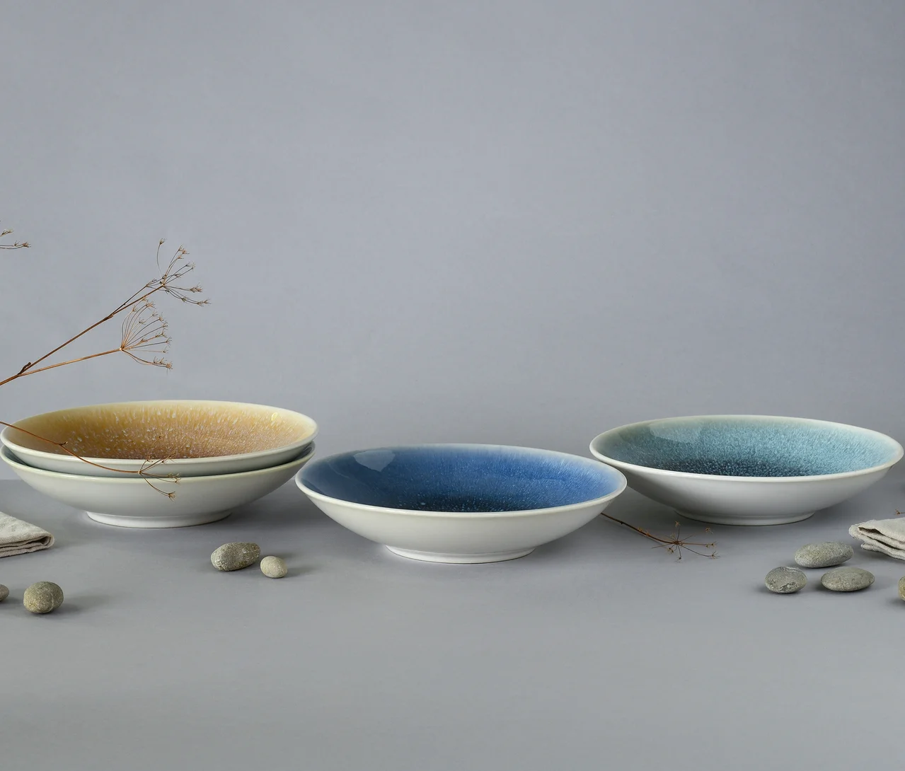 Drei Schalen des creaTable Bowl-Sets »Caldera« mit gesprenkelter Glasur, zwei blaue und eine gelbe, stehen auf einer grauen Oberfläche mit Steinen und getrockneten Pflanzenstielen. Eine gelbe Schale ist auf einer weiteren gestapelt, während eine dunkelblaue und eine hellblaue Schale einzeln platziert sind.