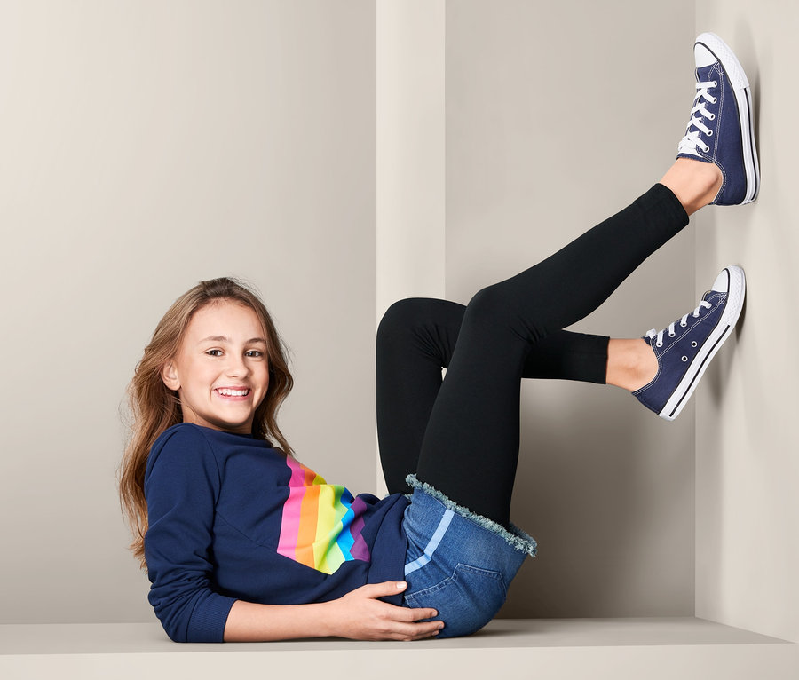 Mädchen liegt und trägt 2 Leggings, Jeansshorts und blaue Turnschuhe.