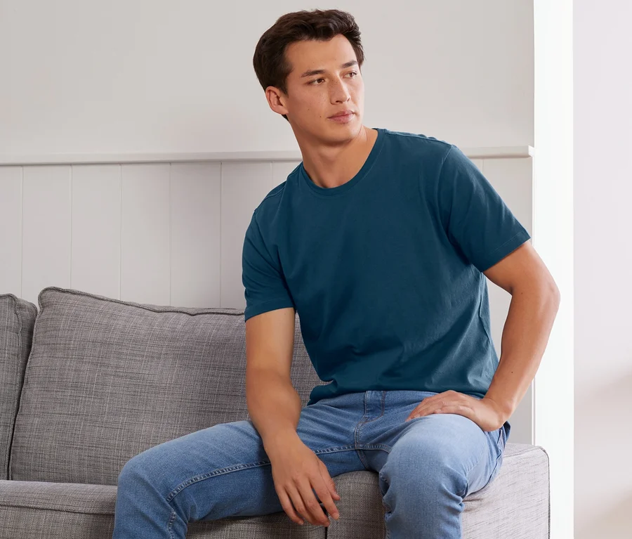 Mann sitzt auf grauem Sofa, trägt ein blaues 2 T-Shirts mit Rundhalsausschnitt.