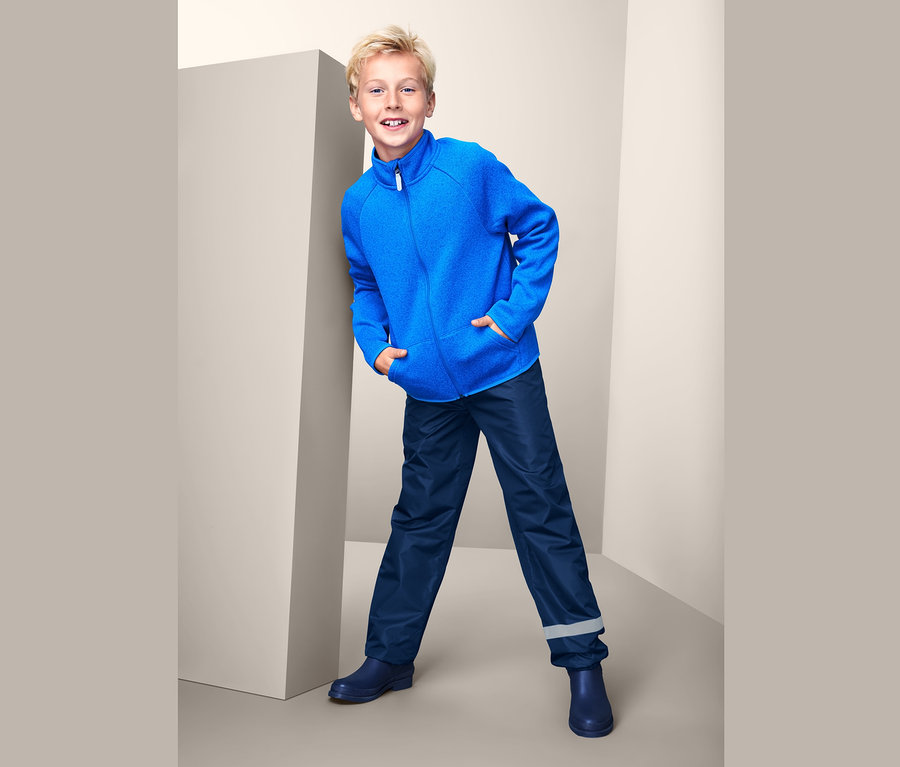 Ein Junge lehnt an einer Wand und trägt eine blaue Jacke und eine Regenhose »Packable«.