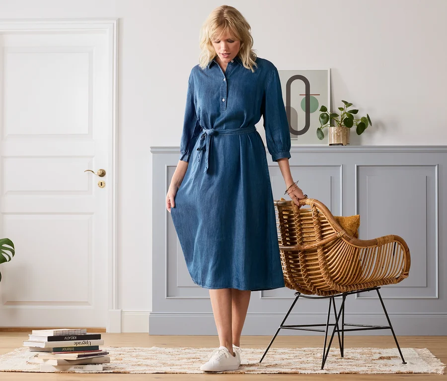 Blonde Frau in einem Midi-Kleid in Denimoptik steht neben einem Rattansessel.