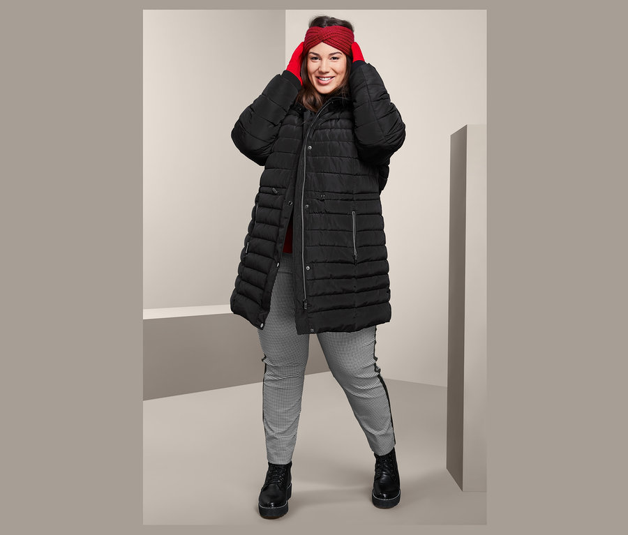 Frau, die ein rotes Strickstirnband richtet, trägt einen schwarzen Steppmantel mit Kapuze und eine knöchellange Stretchhose.