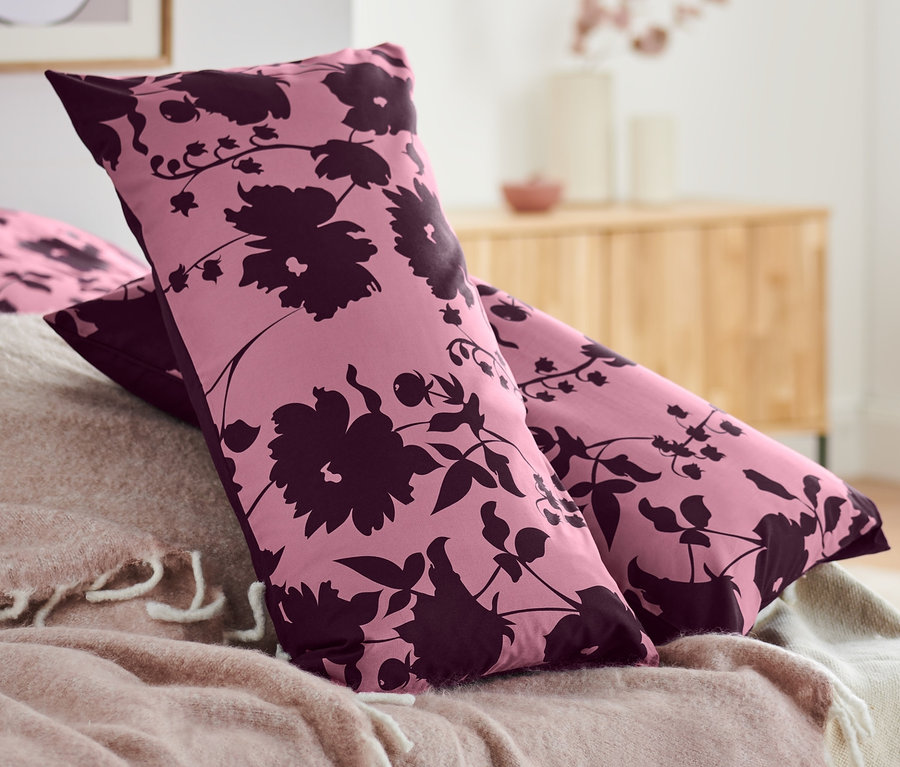 Zwei Renforcé-Kopfkissenbezüge mit Blumenmuster liegen auf einem Bett mit rosa Decke.