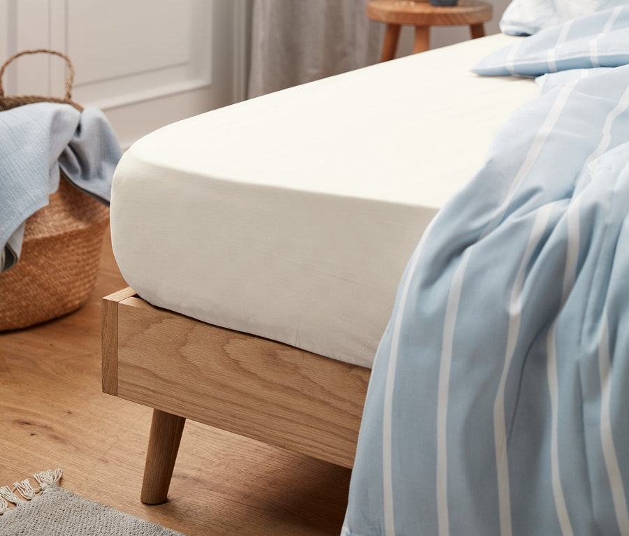 Ein beiges Jersey-Spannbettlaken in Normalgröße ist auf ein Bett mit Holzrahmen aufgezogen. Im Hintergrund befindet sich ein Korb mit blauem Stoff.