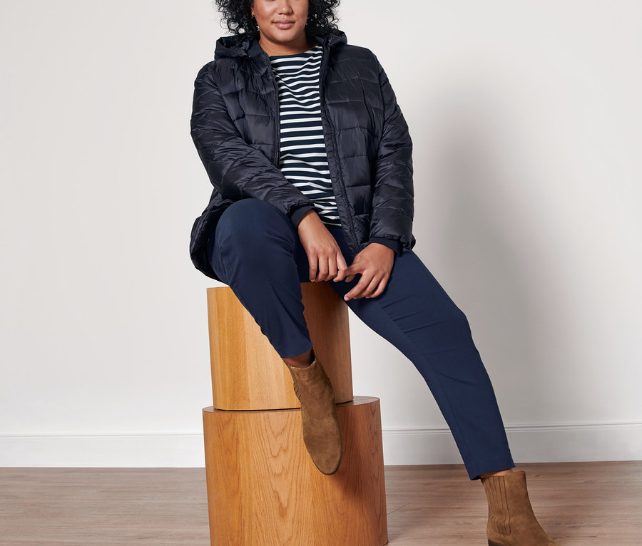 Eine Frau sitzt auf einem Holzhocker und trägt ein blau gestreiftes Langarmshirt, eine dunkelblaue Stretchhose, Stiefeletten aus Veloursleder und eine dunkelblaue Steppjacke mit Kapuze.