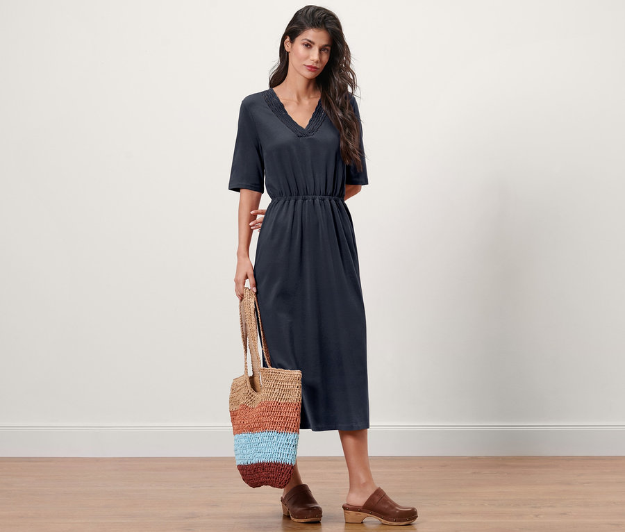 Frau posiert in marineblauem Jerseykleid und braunen Clogs und hält eine gestreifte Tasche.