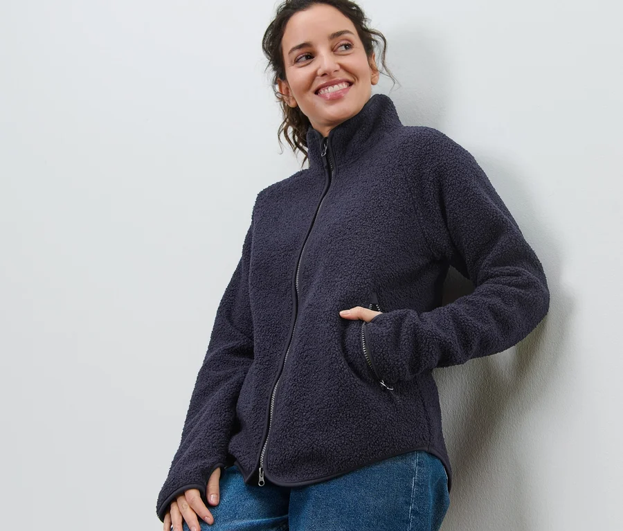 Lächelnde Frau lehnt an einer Wand und trägt eine marineblaue Teddyfleece-Jacke und Jeans.