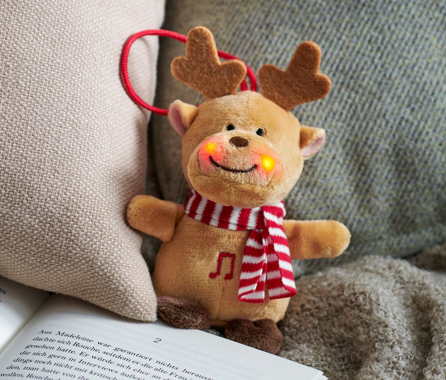 Singendes Mini-Plüschtier Rentier liegt aufgeschlagen neben einem beigen Kissen.