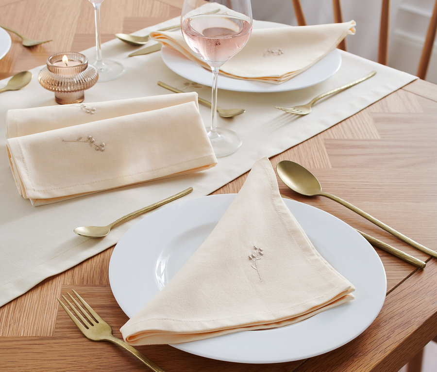 Auf einem Tisch liegen 4 bestickte Servietten, Gläser mit Roséwein, goldenes Besteck und eine Kerze.