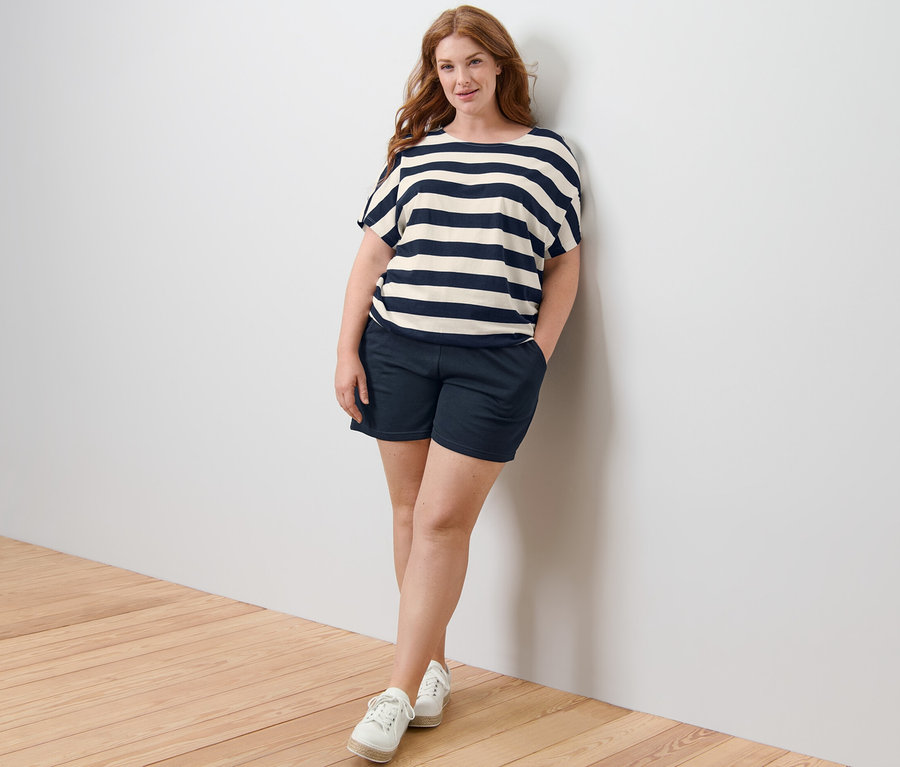 Eine Frau steht an einer Wand, trägt ein gestreiftes Blusenshirt und Sweatshorts.