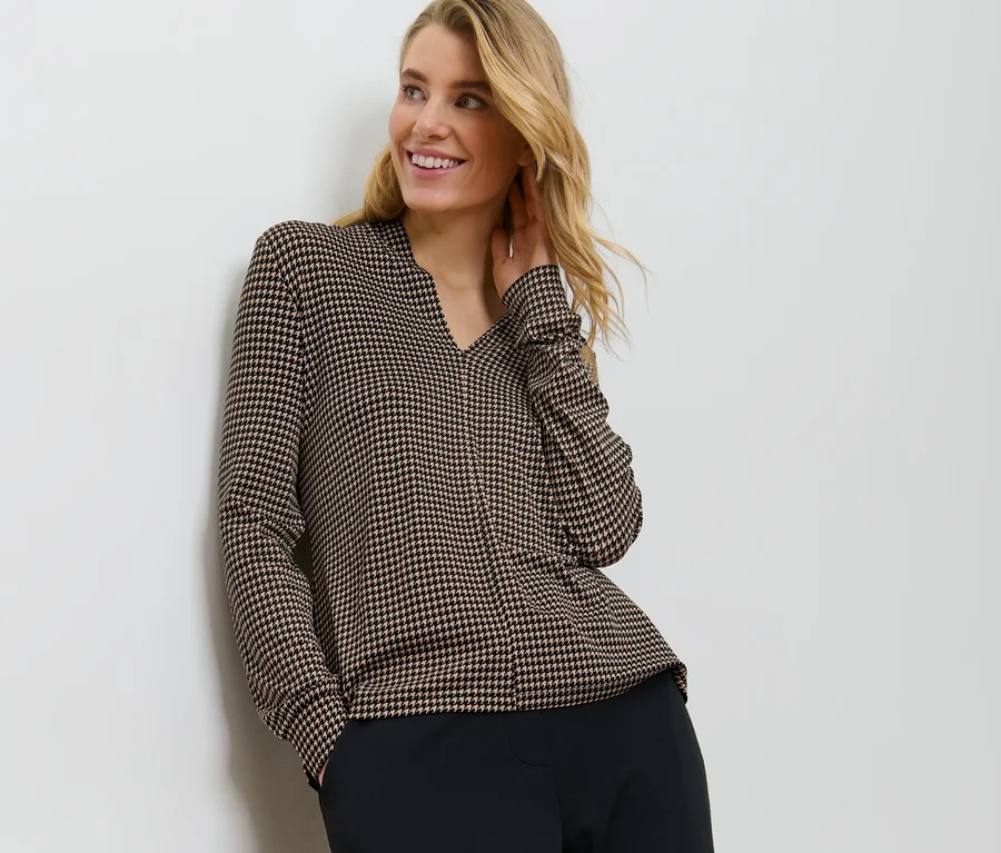 Blonde Frau, an eine Wand gelehnt, trägt ein bedrucktes Blusenshirt mit V-Ausschnitt.