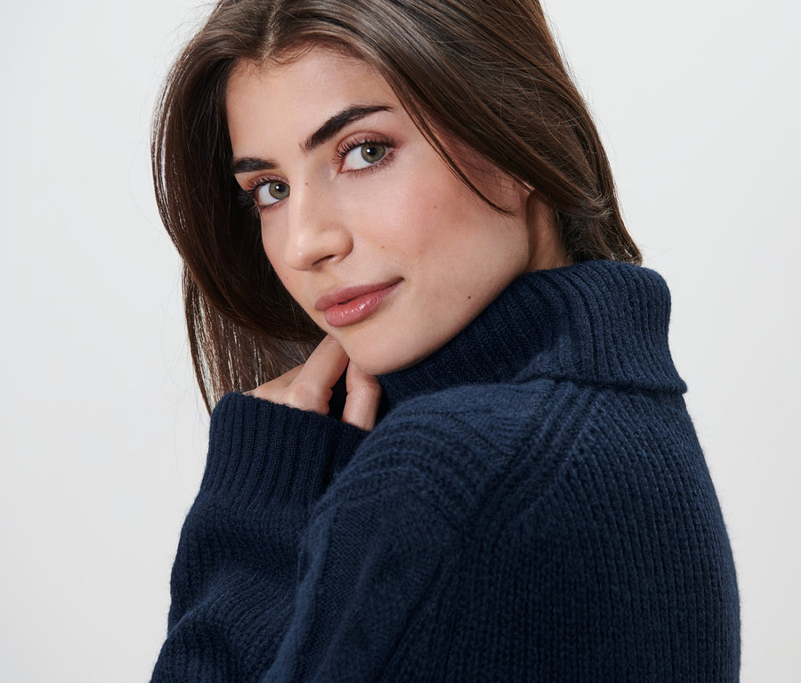 Frau blickt über die Schulter und trägt einen langen Strickpullover mit Zopfmuster.