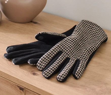Zwei Handschuhe im Materialmix liegen auf einem Holztisch neben einer Vase mit Blumen.