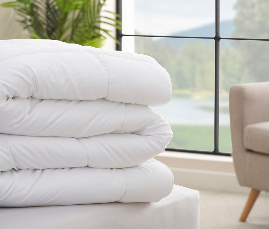 Ein Stapel weißer Home By TEMPUR® Luxe Duo-Bettdecken liegt auf einem Bett neben einem Fenster.