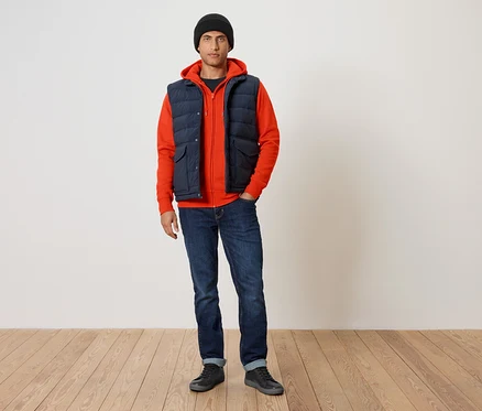 Mann trägt einen orangefarbenen Kapuzenpullover, eine dunkelblaue Steppweste und blaue Jeans.