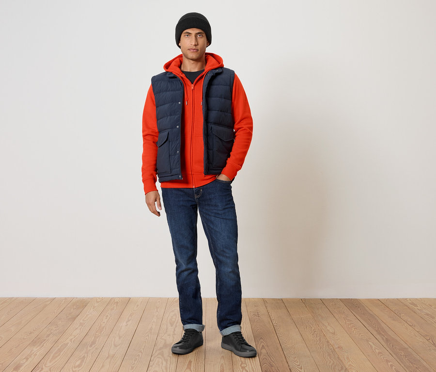 Mann trägt einen orangefarbenen Kapuzenpullover, eine dunkelblaue Steppweste und blaue Jeans.
