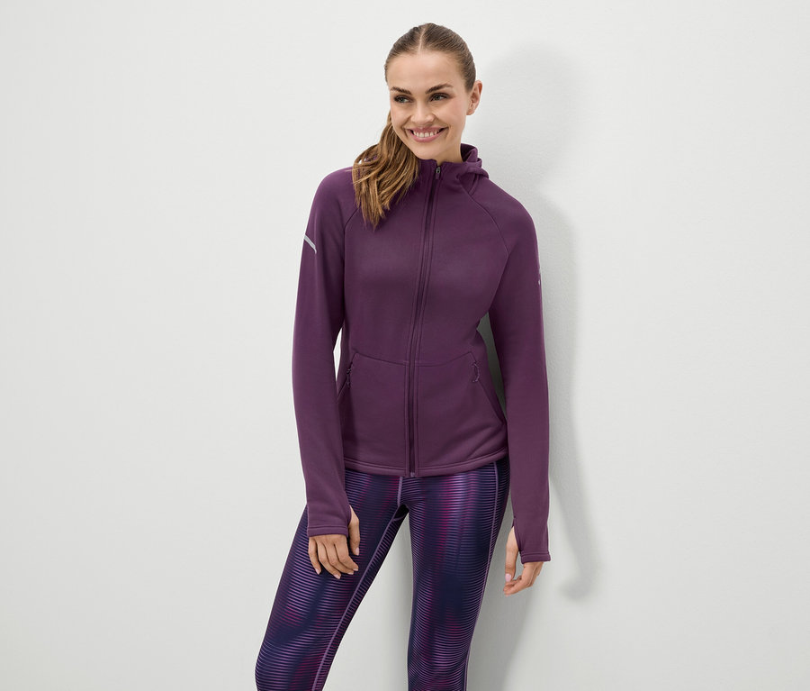 Lächelnde Frau posiert in lila Kapuzenjacke und gemusterten Leggings.