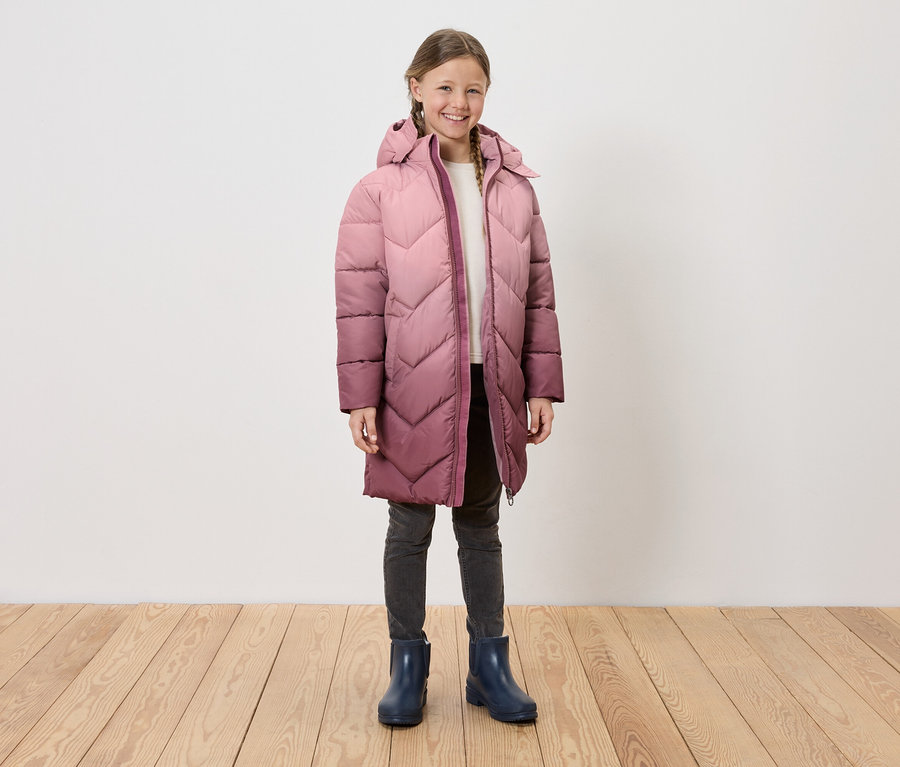 Mädchen in langem, gestepptem Kapuzenmantel, grauen Jeans und blauen Gummistiefeln steht auf Holzboden vor einer weißen Wand.