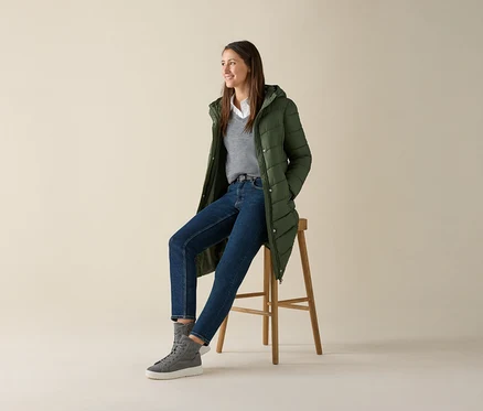 Eine Frau sitzt auf einem Hocker und trägt einen grauen Merino-Feinstrickpullover, Jeans und eine grüne Jacke.