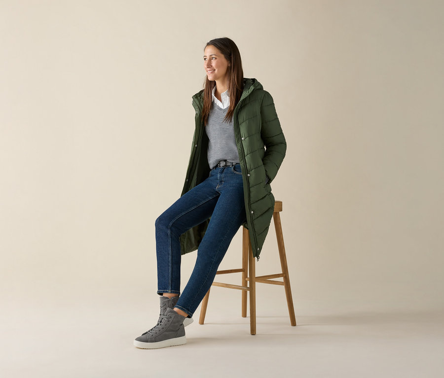 Eine Frau sitzt auf einem Hocker und trägt einen grauen Merino-Feinstrickpullover, Jeans und eine grüne Jacke.