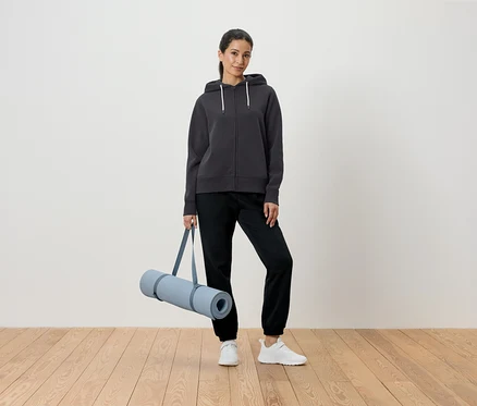 Frau hält Yogamatte in grauem Hoodie und schwarzen Hosen.