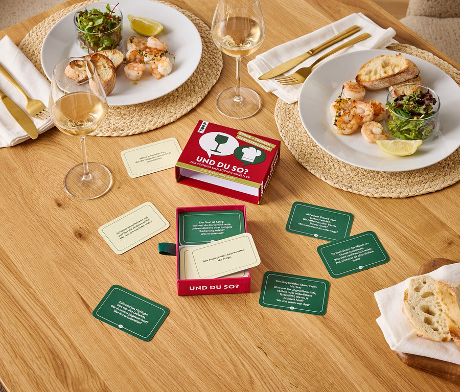 Auf einem Holztisch stehen zwei Teller mit Garnelen, Salat und Brot, zwei Gläser Wein und ein Und du so? Spiel.