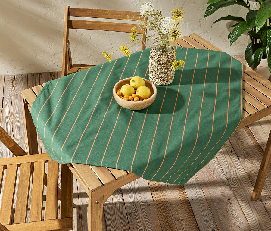 Auf einem Holztisch liegt eine abwischbare Tischdecke, ca. 90 x 90 cm, gestreift. Auf der Tischdecke stehen eine Schale mit Obst und eine Vase mit Blumen.