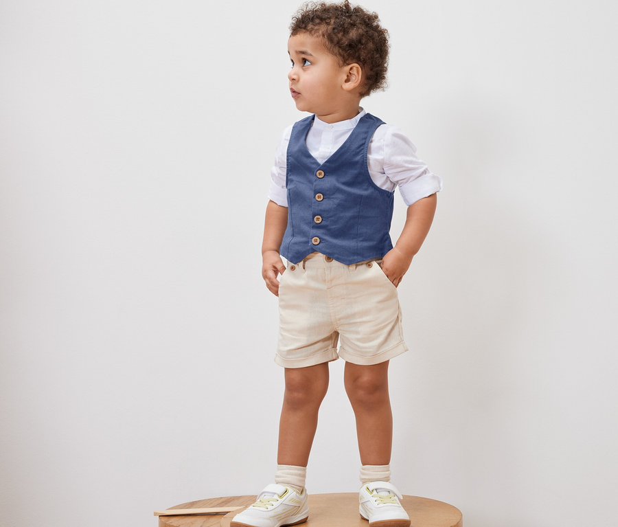 Kleiner Junge steht auf einem Holzhocker und trägt eine Kinder-Weste, ein Hemd, Shorts und Schuhe.