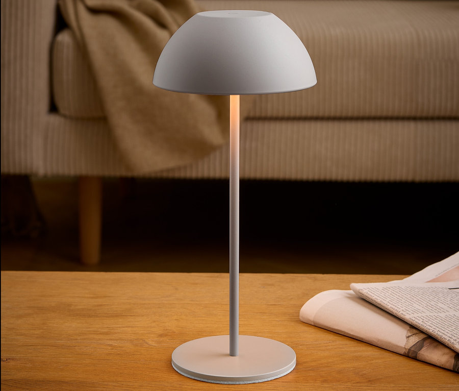 Eine graue Lampe steht neben einer Zeitung auf einem Holztisch. Im Hintergrund befindet sich ein beiges Sofa.