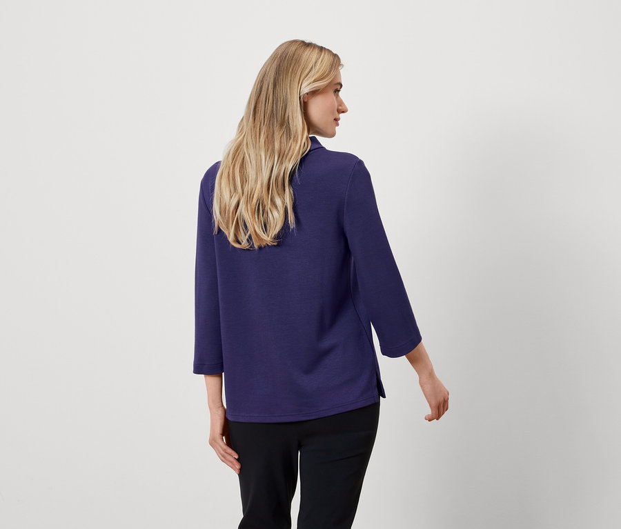 Von hinten sieht man eine Frau mit blonden Haaren, die eine blaue Bluse mit Dreiviertelärmeln und eine schwarze Hose trägt.