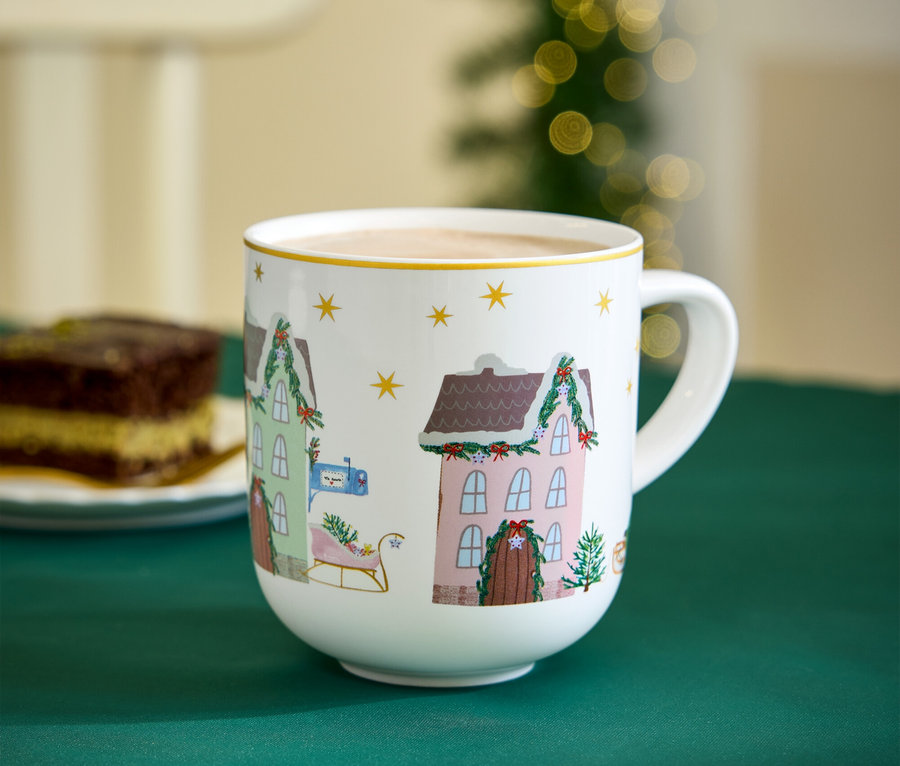 Eine Tasse mit Weihnachtsmotiven steht auf einem grünen Tuch neben einem Stück Kuchen.