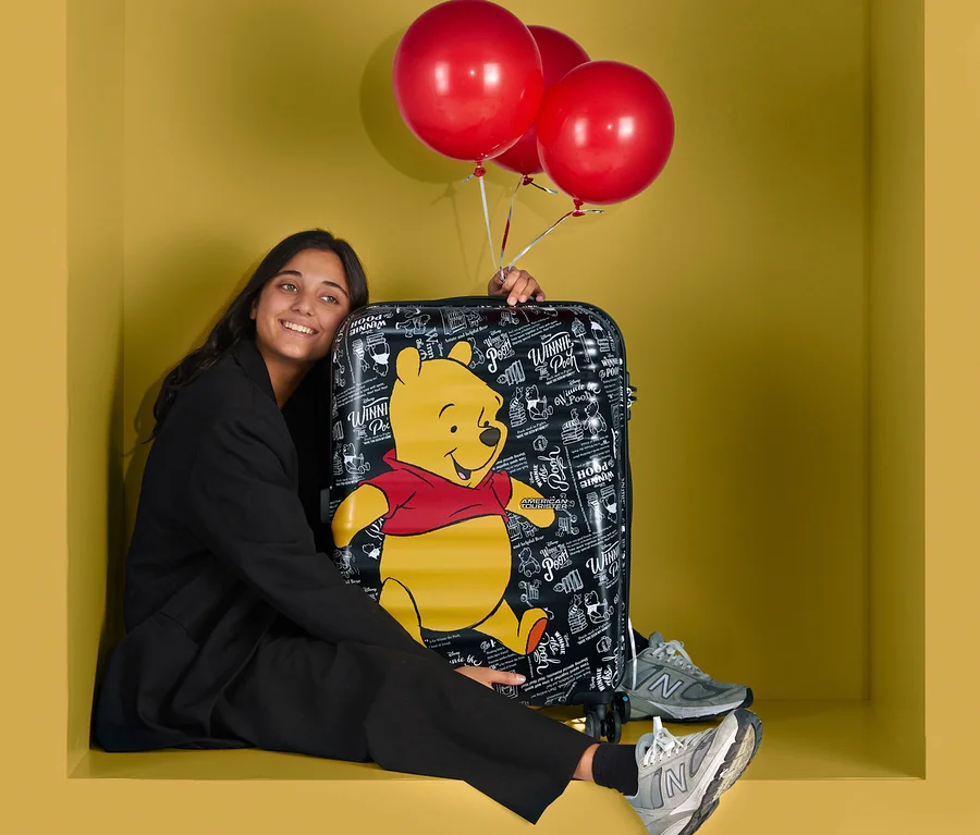 Eine Frau sitzt in einer gelben Box neben einem kleinen American Tourister Wavebreaker »Winnie Poo« und hält drei rote Ballons.