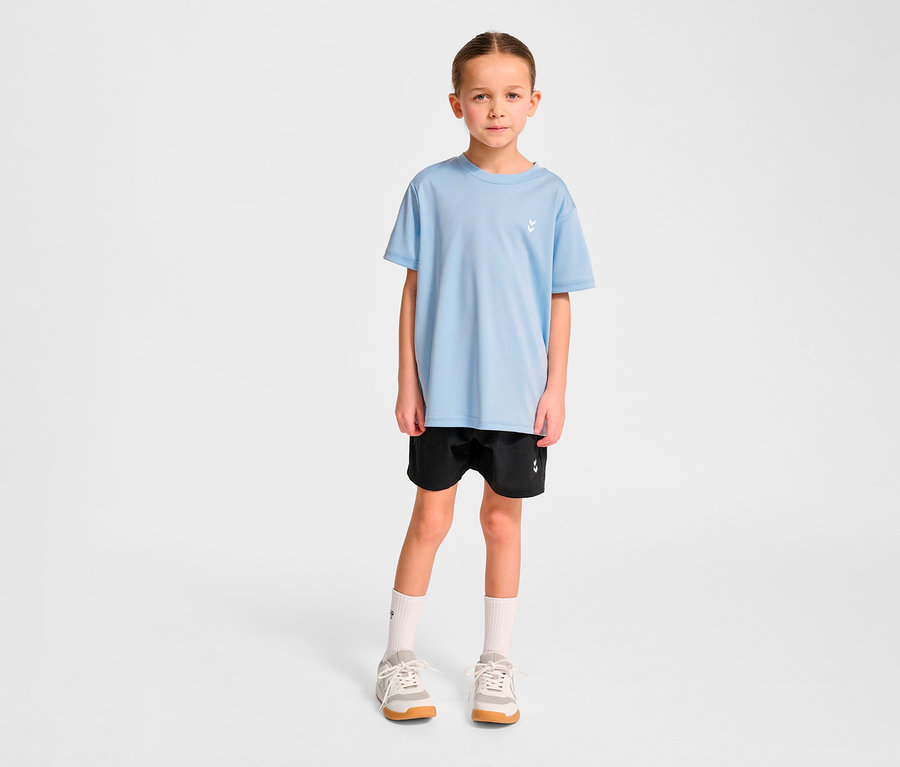 Mädchen steht und trägt ein hellblaues HUMMEL HMLJR pulse Training-T-Shirt, schwarze Shorts, weiße Socken und Turnschuhe.