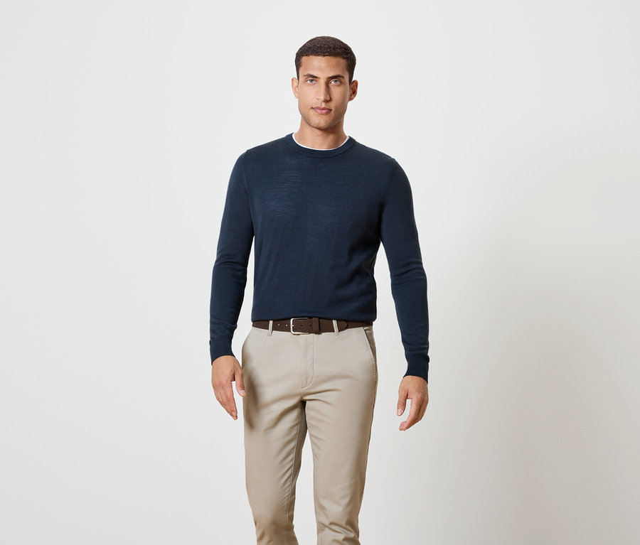 Mann steht und trägt einen dunkelblauen Pullover, einen braunen Gürtel und Khakihosen.