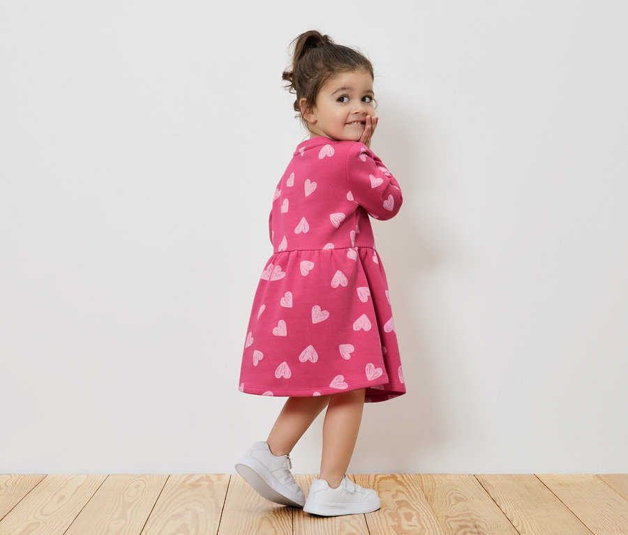 Mädchen in rosa Kleid mit Herzen und weißen Turnschuhen schaut über die Schulter.