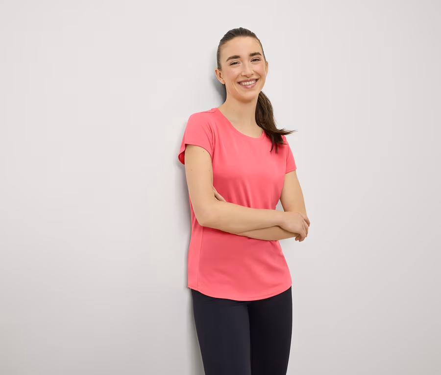 Eine lächelnde Frau mit braunen Haaren im Pferdeschwanz steht mit verschränkten Armen vor der Brust. Sie trägt ein rosa T-Shirt und marineblaue Leggings.