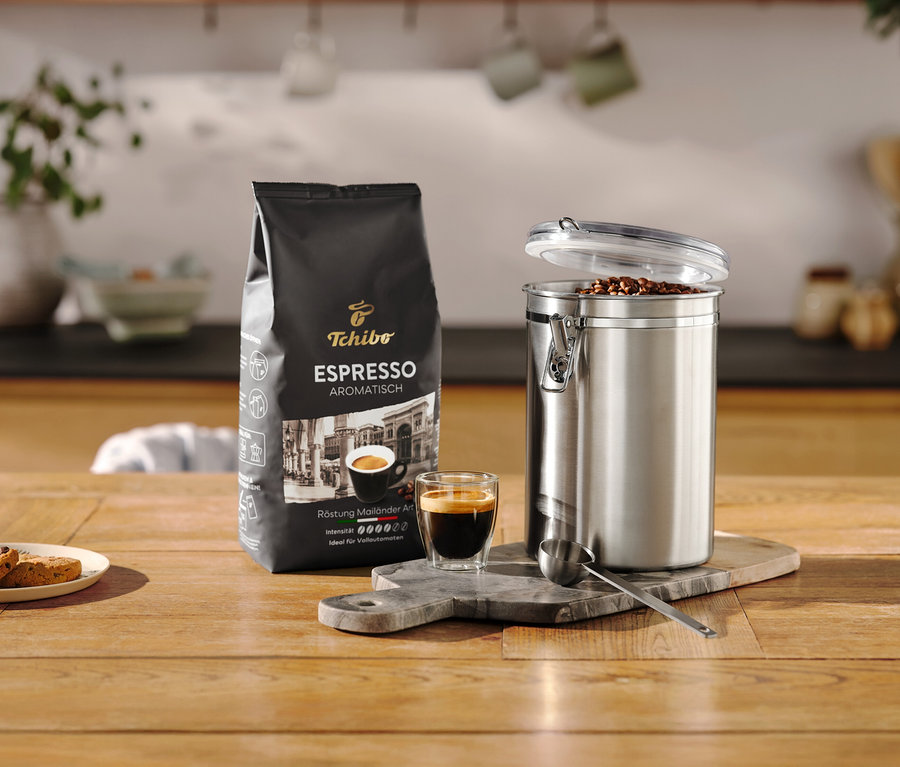 Espressopackung steht neben geöffneter Aromadose aus Edelstahl und Tasse auf einem Holztisch.