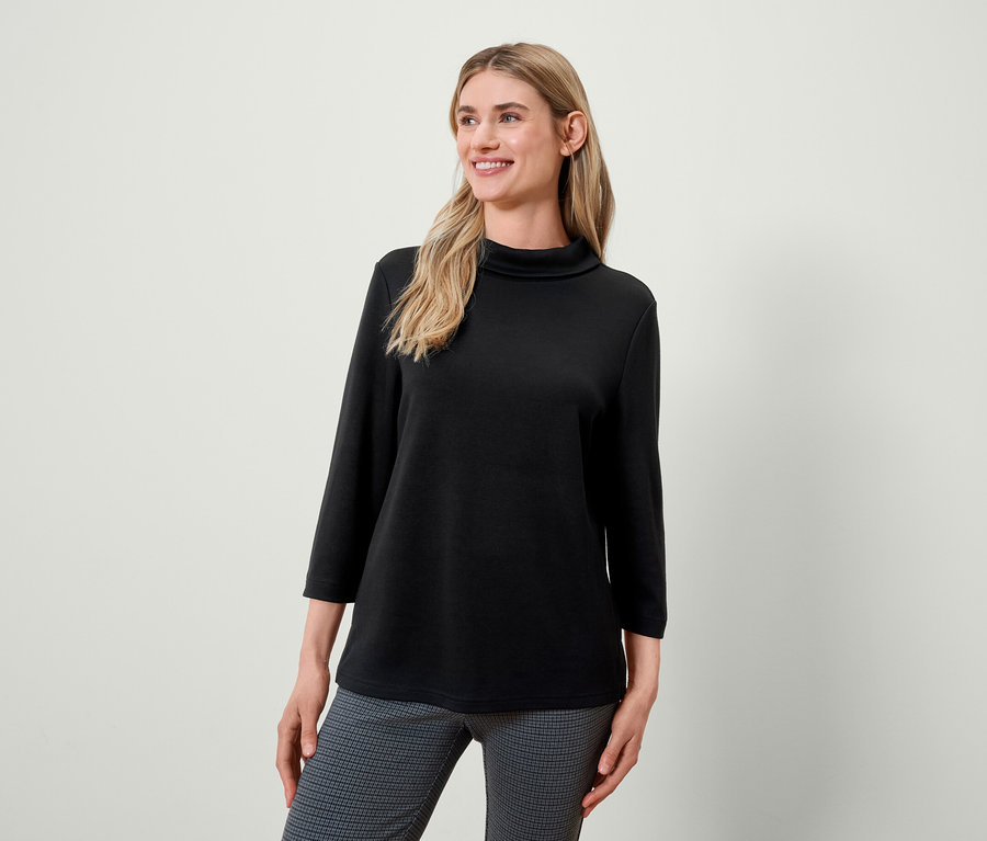 Blonde Frau mit schwarzem Rollkragenpullover und grau gemusterter Hose lächelt und schaut nach links.