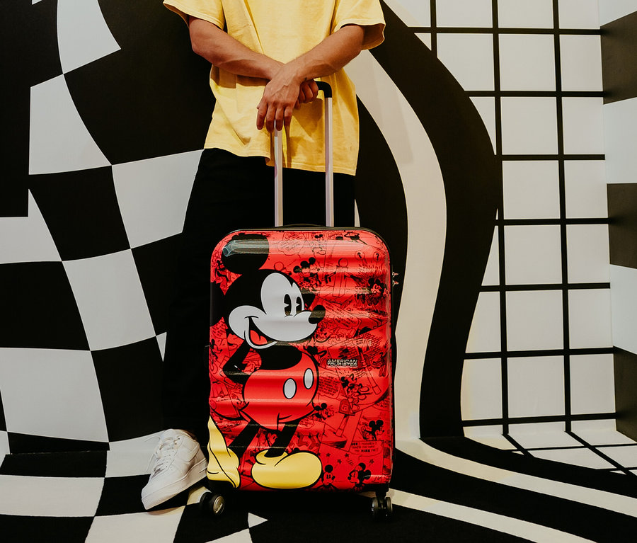 Mann lehnt an einem mittelgroßen American Tourister Wavebreaker »Mickey Mouse« Koffer.